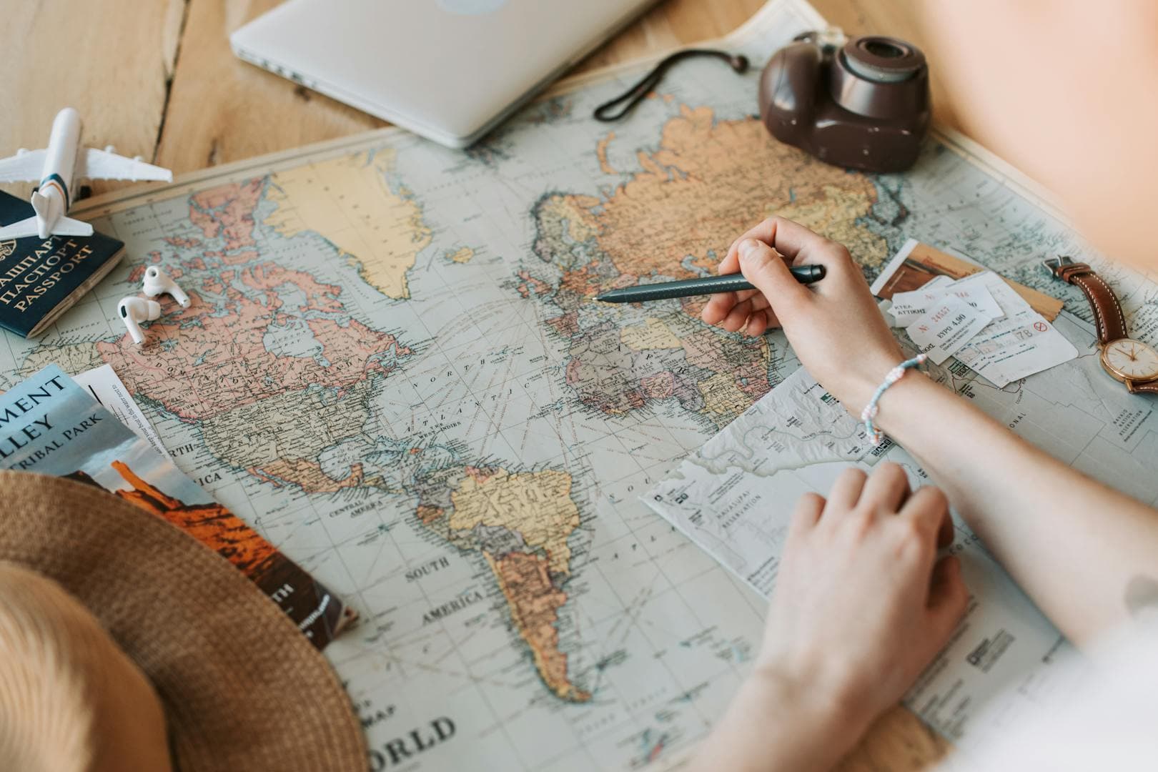 Hands marking a route on a paper world map surrounded by a passport, camera and ticket stubs