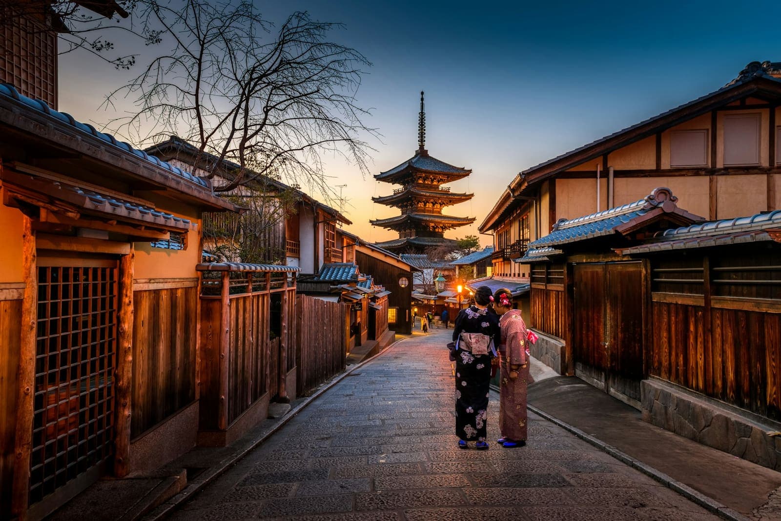 Two people in kimono on a stone lane of wooden machiya houses below a five-storey pagoda at sunset in Kyoto