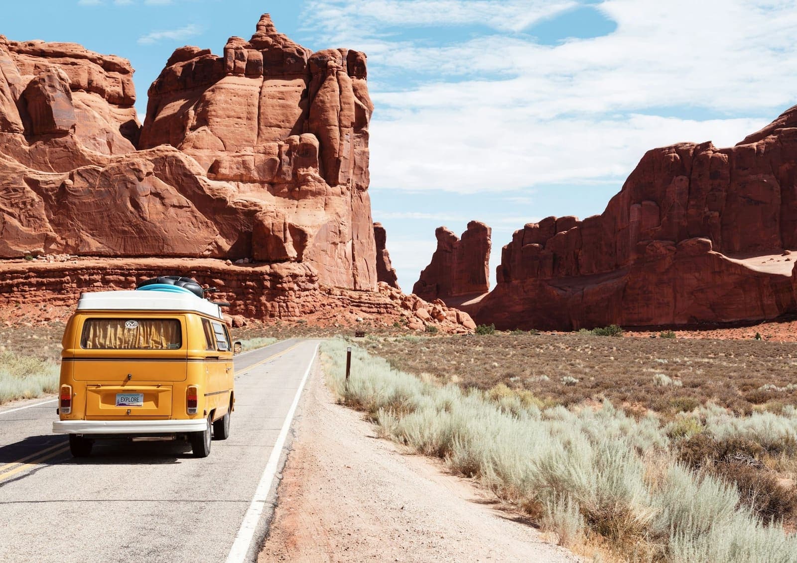 A yellow vintage camper van on a two-lane road running past towering red sandstone fins in the desert