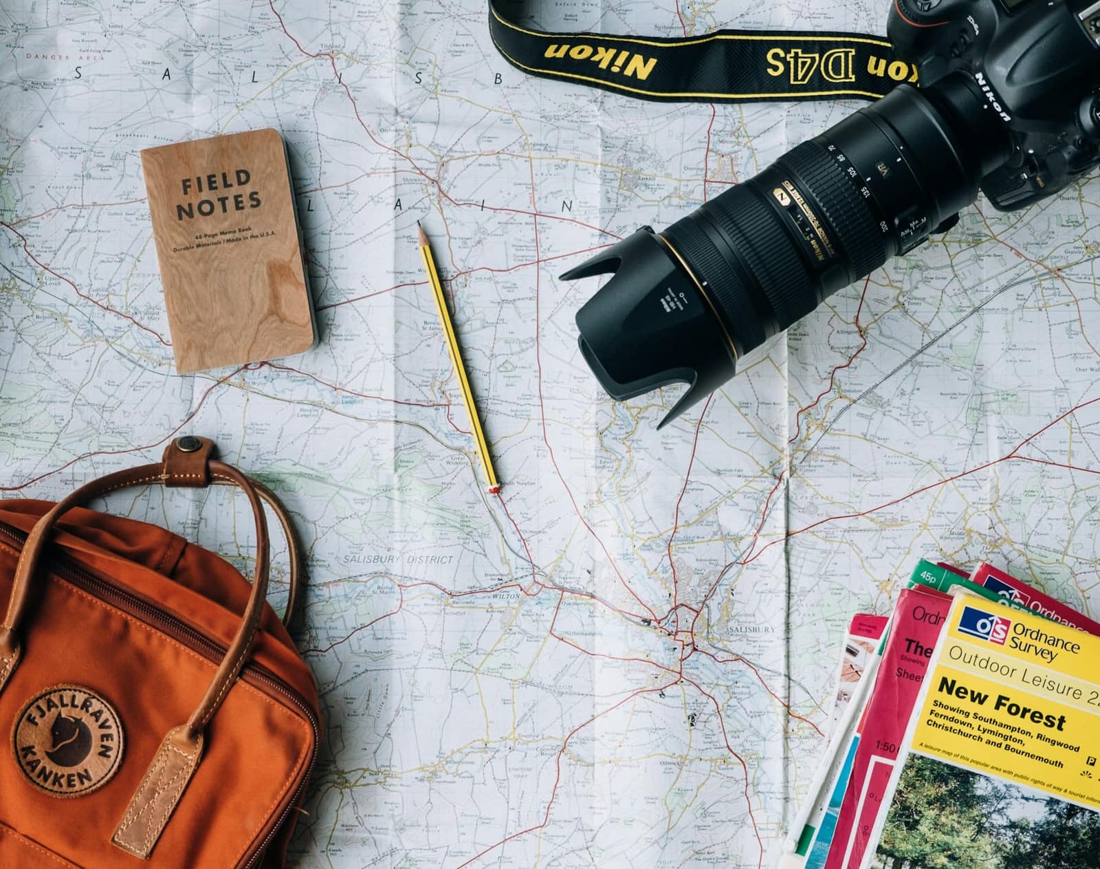 A folded paper map laid flat with a camera, a notebook, a pencil, a rucksack and guide leaflets on top