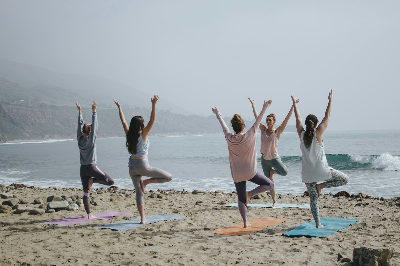 Five people holding tree pose on yoga mats on a sandy beach facing a misty coastline