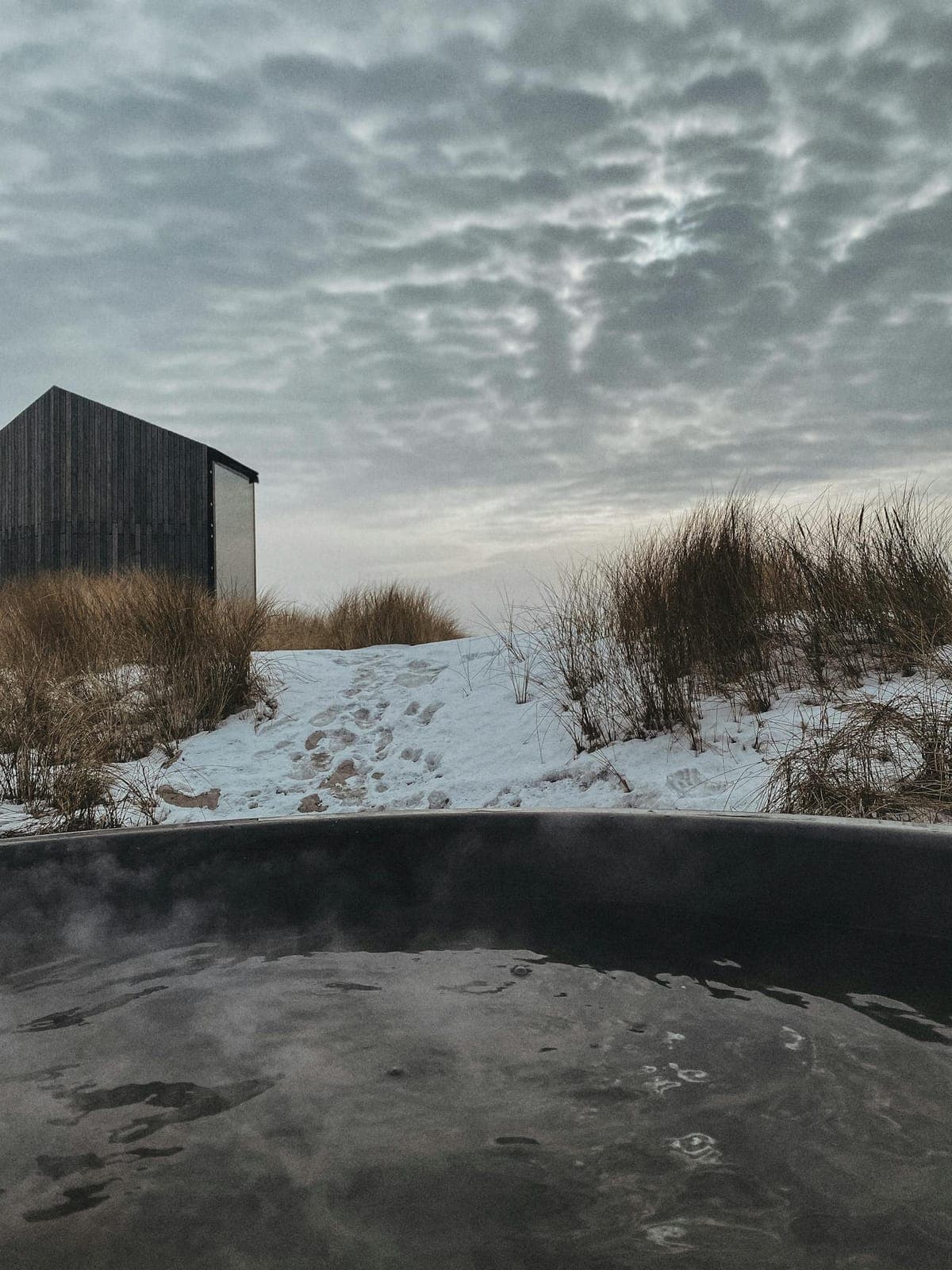 Steam drifting off an outdoor hot tub beside a dark timber cabin in snow-dusted dune grass