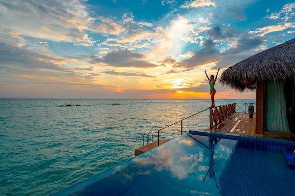 A person standing arms raised on the deck of a thatched overwater villa at sunset