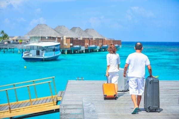 Two travellers wheeling suitcases along a jetty toward thatched overwater bungalows