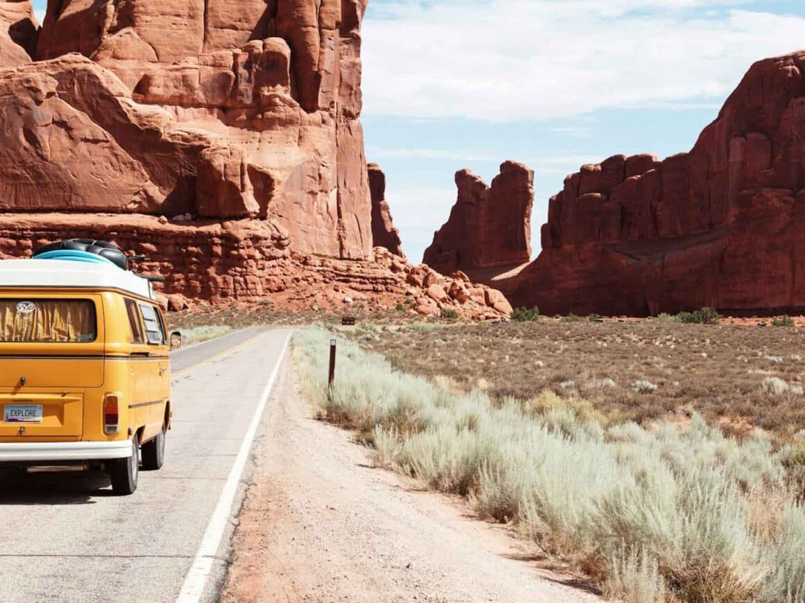 A yellow vintage camper van on a two-lane road running past towering red sandstone fins in the desert